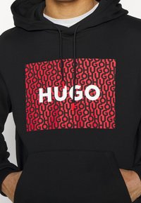 Černá mikina s kapucí se výraznou červenou grafikou s opakujícím se textem "HUGO" a "GO", vyrobená z měkké tkaniny, s přední kapsou a stažitelnou kapucí.