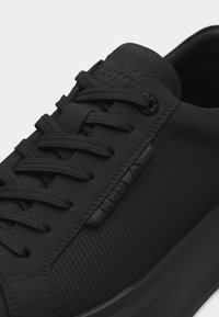 Κοντινό πλάνο ενός μαύρου παπουτσιού Replay με υφασμάτινη υφή, μαύρα κορδόνια και μαύρη πλαϊνή ετικέτα που εμφανίζει το λογότυπο Replay.