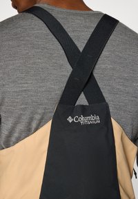 Mustard ja musta Columbia Titanium kott reguleeritavate mustade rihmadega, millel on sile tekstuur ja logode detail. Kandmisel halli särke.
