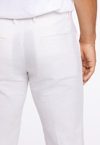 Lněné kalhoty světle béžové barvy s hladkou texturou. Mají dvě zadní kapsy a střih na míru. Na látce jsou viditelné mírné záhyby.