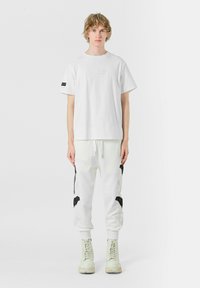 Camiseta de algodón blanca con un parche rectangular en el frente; combinada con pantalones joggers blancos que presentan diseños de pájaros bordados en negro y cremalleras.