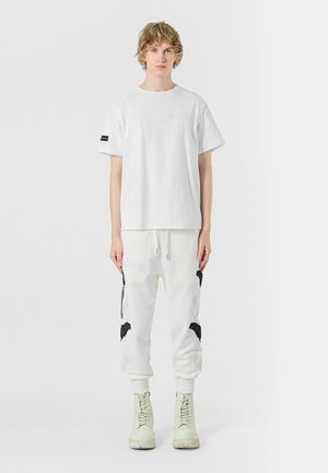 Camiseta de algodón blanca con un parche rectangular en el frente; combinada con pantalones joggers blancos que presentan diseños de pájaros bordados en negro y cremalleras.