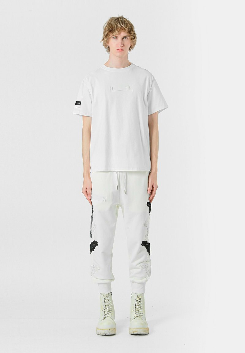Camiseta de algodón blanca con un parche rectangular en el frente; combinada con pantalones joggers blancos que presentan diseños de pájaros bordados en negro y cremalleras.