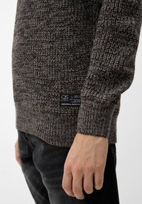 Stickad tröja i mörkgrå nyanser, med ett strukturerat mönster och en etikett längst ner till vänster. Långa ärmar och ribbad manschettdetalj.