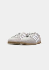 Pelēki zamšādas kedas ar baltām svītrām, bēša gumijas zole, noapaļota purngalu forma un aukliņu aizdare. Minimālisma stils bez papildu detaļām.