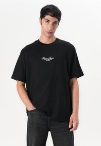 T-shirt noir en coton avec un col rond, des manches courtes et un logo brodé blanc sur la poitrine. Coupe décontractée, longueur standard.