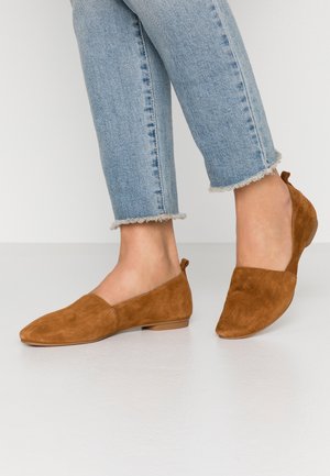 Jean bleu clair à ourlet effiloché associé à des chaussures plates en daim marron sur un sol blanc et un fond clair uni.