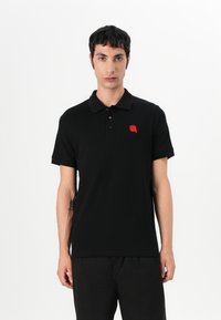 Μαύρο πόλο μπλουζάκι από βαμβάκι με κοντό μανίκι, γιακά, πλάγιο κλείσιμο με τρία κουμπιά και κόκκινο κεντημένο λογότυπο στην αριστερή θήκη του στήθους.