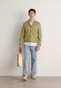 Olijfgroene, knoopsluiting jas met een kraag, gecombineerd met een witte shirt en lichtblauwe spijkerbroek. Houdt een crèmekleurige tas met bruine accenten vast.
