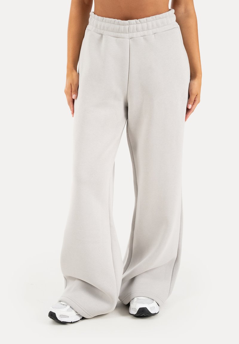 Pantalon de survêtement gris clair à jambes larges avec taille élastique porté par une personne debout, associé à des chaussures de sport blanches.