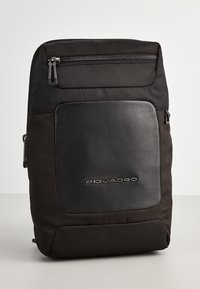 Zaino nero realizzato in tessuto e pelle. Presenta una tasca frontale con zip e un logo del marchio in rilievo. Texture liscia con un design strutturato.