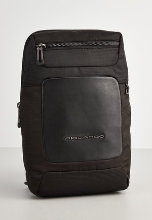 Zaino nero realizzato in tessuto e pelle. Presenta una tasca frontale con zip e un logo del marchio in rilievo. Texture liscia con un design strutturato.