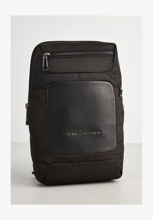 Zaino nero realizzato in tessuto e pelle. Presenta una tasca frontale con zip e un logo del marchio in rilievo. Texture liscia con un design strutturato.