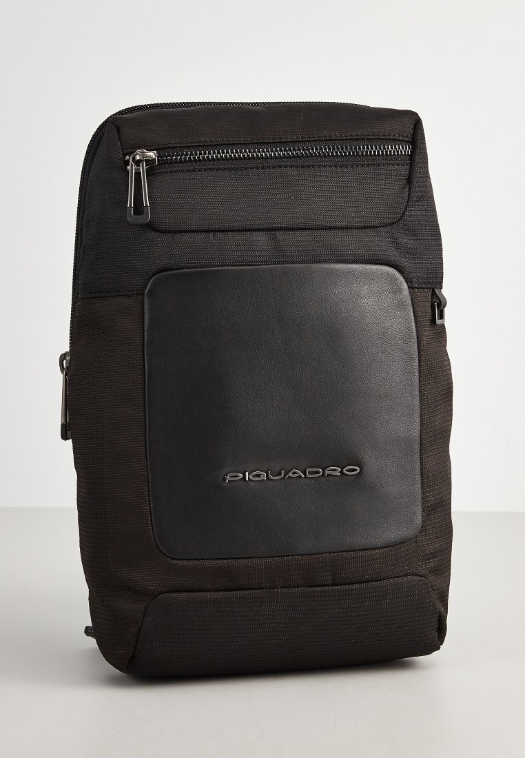 Zaino nero realizzato in tessuto e pelle. Presenta una tasca frontale con zip e un logo del marchio in rilievo. Texture liscia con un design strutturato.
