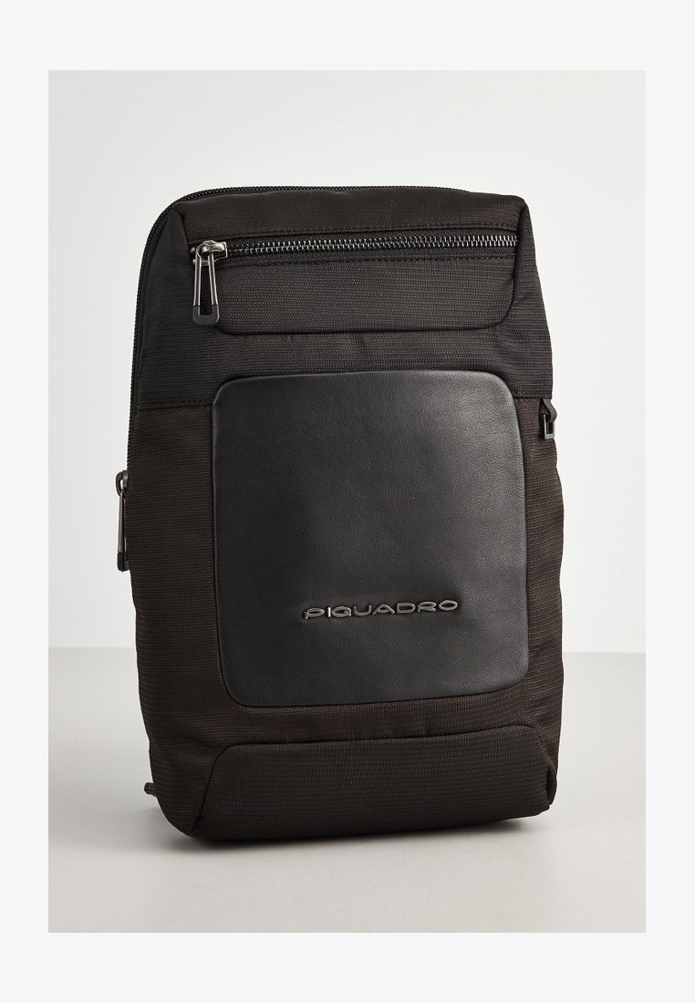 Zaino nero realizzato in tessuto e pelle. Presenta una tasca frontale con zip e un logo del marchio in rilievo. Texture liscia con un design strutturato.