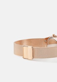 Rose gold mesh watch band with a polished metal clasp. The design features a fine mesh texture and an angular watch face.