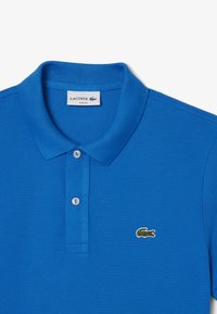Camisa polo azul hecha de tejido texturizado, con un collar clásico, dos botones y un pequeño logo de cocodrilo verde en el pecho.