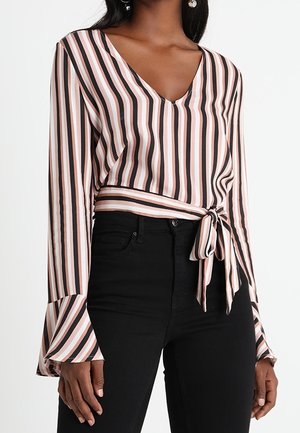 Mujer que lleva una blusa de rayas de manga larga con escote en V, puños acampanados y un lazo lateral, combinada con pantalones negros de cintura alta.