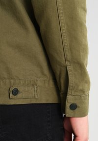 Manche de veste olive avec un bouton et un détail de poignet. Texture de tissu lisse avec des coutures visibles le long de la couture.