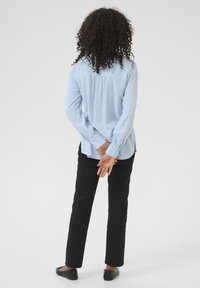 Himmelblaues Langarmhemd mit Rückenfalte, kombiniert mit schwarzen Slim-Fit-Hosen und schwarzen Ballerinas. Lockige Haare sind oberhalb des Hemdkragens sichtbar.