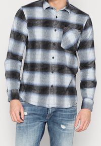 Hombre vestido con una camisa de cuadros azul, blanca y negra con un bolsillo frontal, combinada con jeans desgastados de color azul claro.