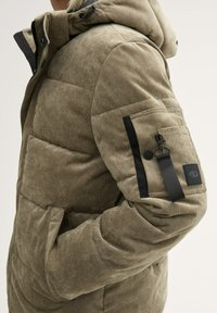 Geplüschte Jacke aus olivgrünem Baumwollcord mit strukturiertem Finish. Sie verfügt über eine Kapuze, schwarze Akzente, Seitentaschen und eine Reißverschlusstasche am Ärmel.