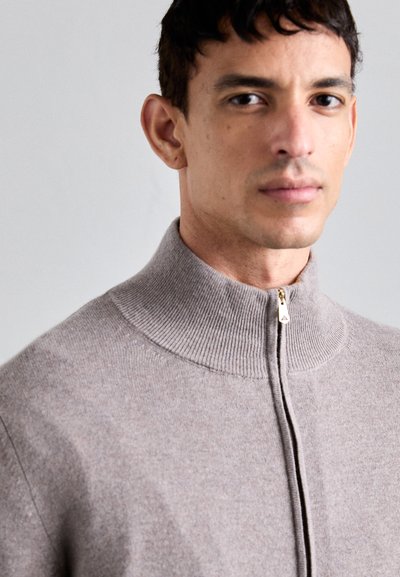 Svetlo siva, zip-up jopica iz mehke pletenine. Ima visok ovratnik in subtilno rebrasto teksturo, s podrobnostjo z zlatim zadrgo.