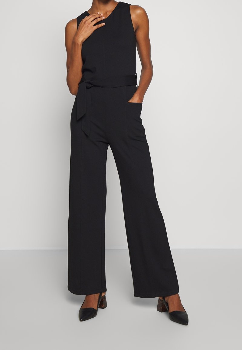 Combinaison sans manches noire avec des jambes larges, ceinture à nouer à la taille et poches avant ; fabriquée en tissu lisse avec un minimum de détails de coutures.