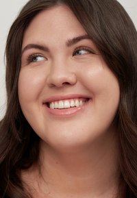 Gros plan d'une personne souriante avec des cheveux noirs mi-longs, une peau claire, un maquillage naturel et une couleur de lèvres claire sur un fond neutre.