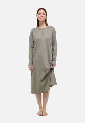 Femme debout pieds nus portant une robe midi boutonnée manches longues de couleur vert olive, tenant un pli de la jupe avec sa main droite.