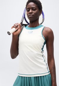 Γυναίκα που κρατά ρακέτα του τένις πάνω στον ώμο της, φορώντας λευκό αμάνικο τοπ με τιρκουάζ περιγράμματα και φούστα με πιέτες στο ίδιο τιρκουάζ χρώμα.