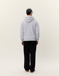 Felpa grigia chiara con cappuccio e cordoncino, dotata di tasca a forma di canguro, abbinata a pantaloni neri e un cappello scuro, vista da dietro.