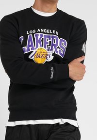 Uomo che indossa una felpa nera dei Los Angeles Lakers con logo viola e bianco, braccia incrociate, sovrapposta a una maglietta bianca e pantaloni neri.