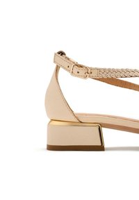 Sandal con tacco blocco beige con cinturino alla caviglia intrecciato. Presenta un accento dorato lucido sul tacco e una trama in pelle liscia.