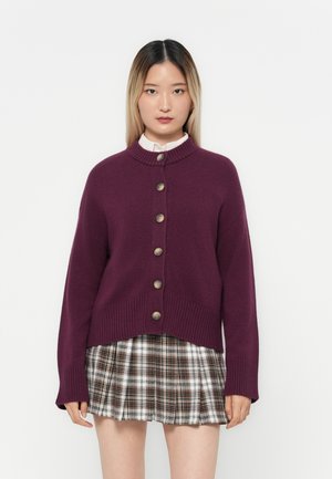 Cardigan en tricot violet avec un col haut, fermeture à boutons sur le devant et ourlet côtelé. Porté par-dessus une jupe à carreaux, avec des motifs de checks variés.