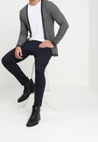 Grå cardigan med svart kant över en vit t-shirt, tillsammans med marinblå byxor och svarta snörstövlar, sittande på en vit pall.