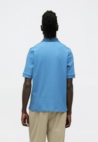 Πόλο μπλούζα με κοντά μανίκια σε ανοιχτό μπλε, υφασμάτινη με υφή, με γιακά και διπλωτό σχέδιο. Η πίσω όψη δείχνει μια άνετη εφαρμογή.