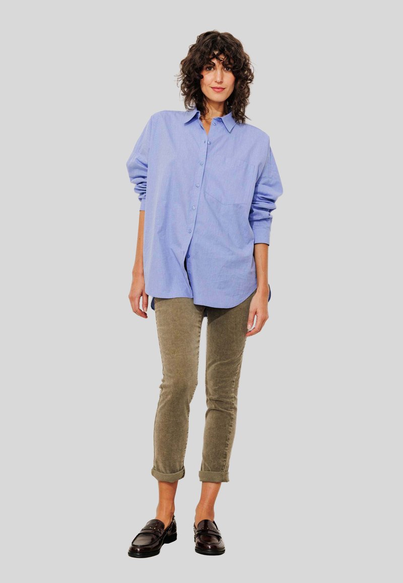 Femme aux cheveux bouclés portant une chemise oversize bleu clair à boutons, un pantalon en velours côtelé olive retroussé et des mocassins noirs se tenant devant un fond gris.