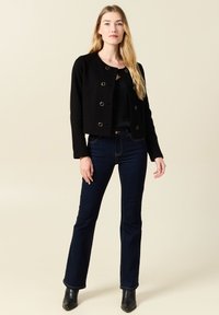 Zwarte cropped jas met ronde hals en goudkleurige drukknoopsluitingen, gecombineerd met donkere denim flared jeans en zwarte enkellaarsjes.