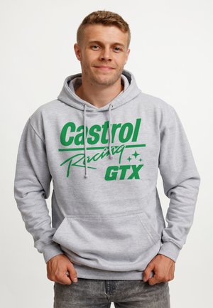 Junger Mann in grauem Kapuzenpullover mit grünem "Castrol Racing GTX"-Text, steht mit den Händen in Taillennähe und lächelt leicht vor einem schlichten Hintergrund.