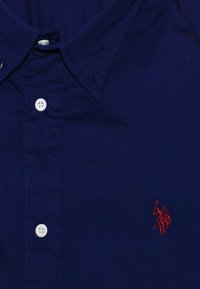 Tummansininen puuvillasta valmistettu napillinen paita, jossa on terävä kaulus, valkoiset napit ja vasemmassa rinnassa pieni punainen brodeerattu logo.