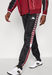 Czarne, zwężane spodnie dresowe z białymi i czerwonymi bocznymi paskami z logo Kappa. Matowe wykończenie, elastyczna talia i ściągane nogawki.