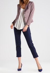 Femme portant un blazer mauve, un chemisier plissé blanc, un pantalon cropped bleu marine et des talons hauts bleu marine, posant devant un fond clair uni.