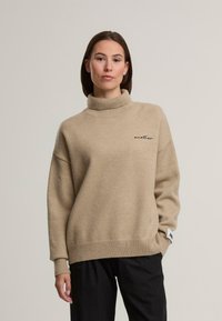 Beżowy sweter z golfem wykonany z miękkiego materiału. Charakteryzuje się luźnym krojem, ściągaczami przy rękawach oraz haftowanym napisem "another" na klatce piersiowej.