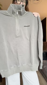 Sweatshirt cinza de fleece com fecho zip, colarinho alto, barra e punhos em malha. Apresenta um logo preto "PEGADOR" na frente. Textura macia.