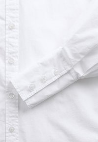 Bela bombažna srajca z manšetom na štiri gumbe, preprostega dizajna in gladke teksture. Ovratnik je viden na levi strani.