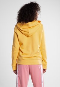 Gul hoodie med mjuk textur, utrustad med en rymlig huva och ribbstickad fåll. Bärs med rosa byxor som har vita sidostrådar.