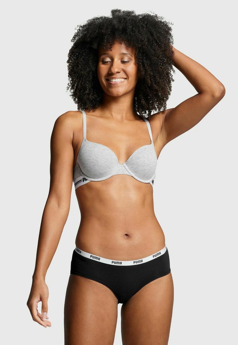 Lächelnde Frau mit lockigem Haar, die einen hellgrauen BH und schwarze Puma-Unterwäsche trägt, posiert mit einer Hand hinter ihrem Kopf.