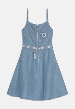 Robe en denim bleu clair sans manches avec fermeture éclair frontale, poche poitrine affichant le logo "cK", et ceinture blanche de marque Calvin Klein à la taille.