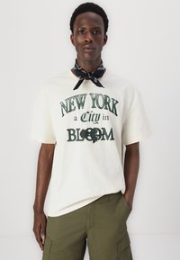 Creme t-shirt med grøn trykt tekst "NEW YORK en By i BLOOM," prydet med et blomsterdesign. Kombineret med olivengrønne cargo shorts og et sort bandana.